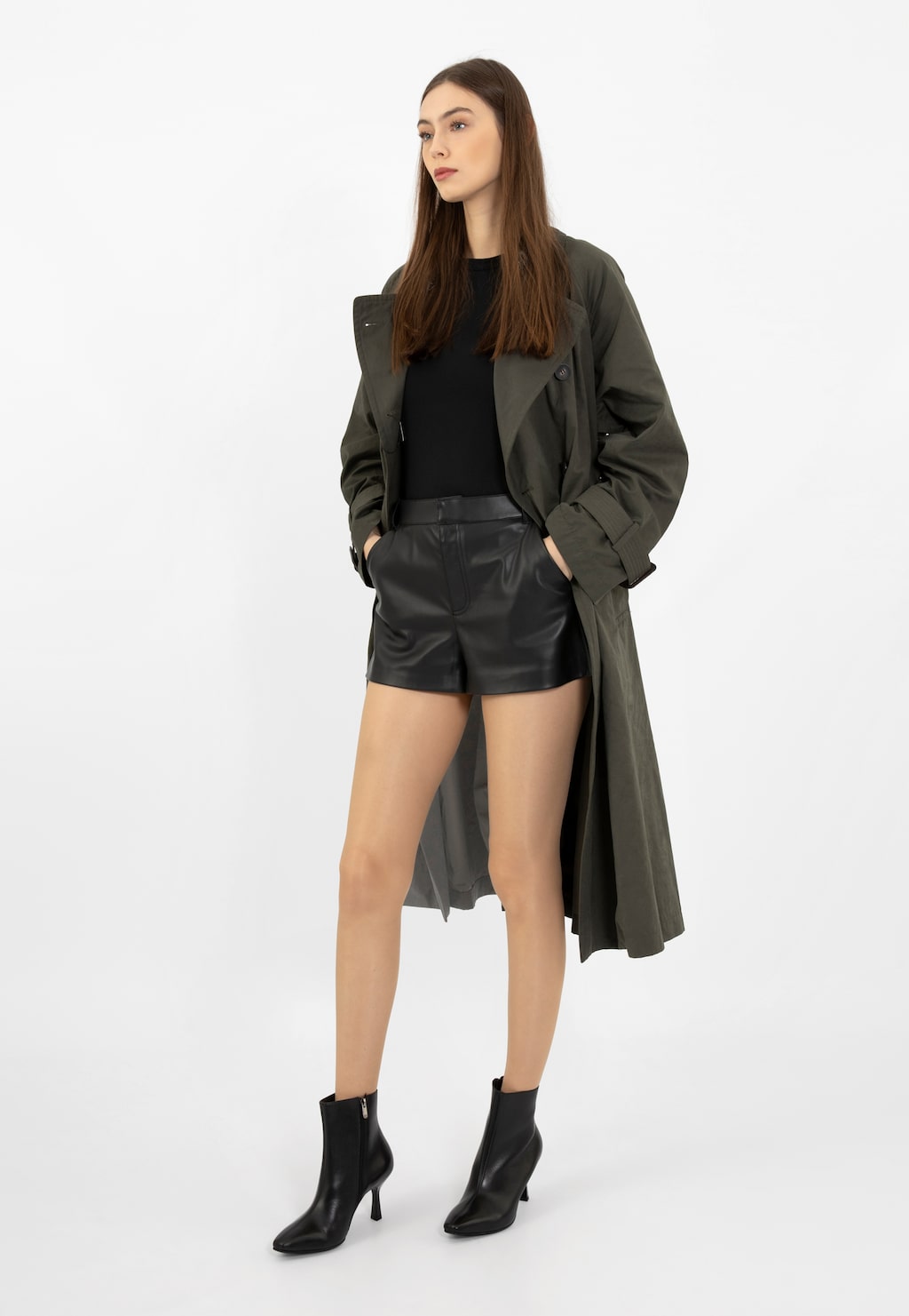 Modelka w stylizacji z czarnymi botkami Wojas 55207-51 na szpilce, czarnym topem, skórzanymi szortami i oliwkowym płaszczem. Elegancki look podkreślający smukłą sylwetkę.