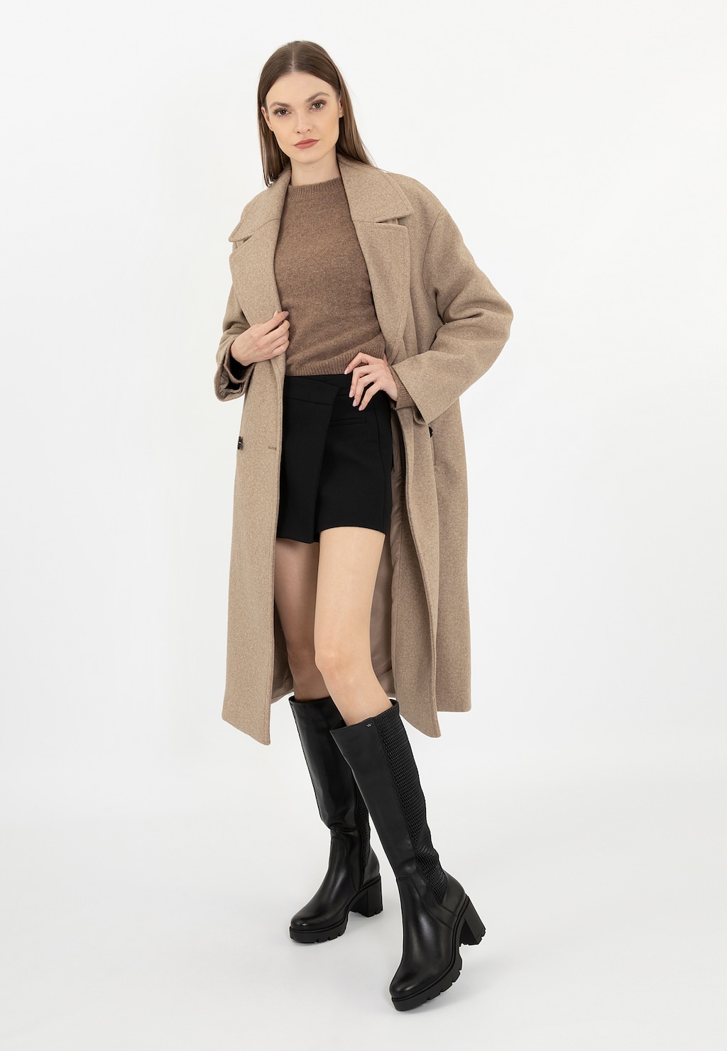 Modelka w beżowym płaszczu i brązowym swetrze prezentująca czarne kozaki damskie Wojas model 71048-81 na szerokim obcasie. Buty mają elegancki wygląd z grubą podeszwą.