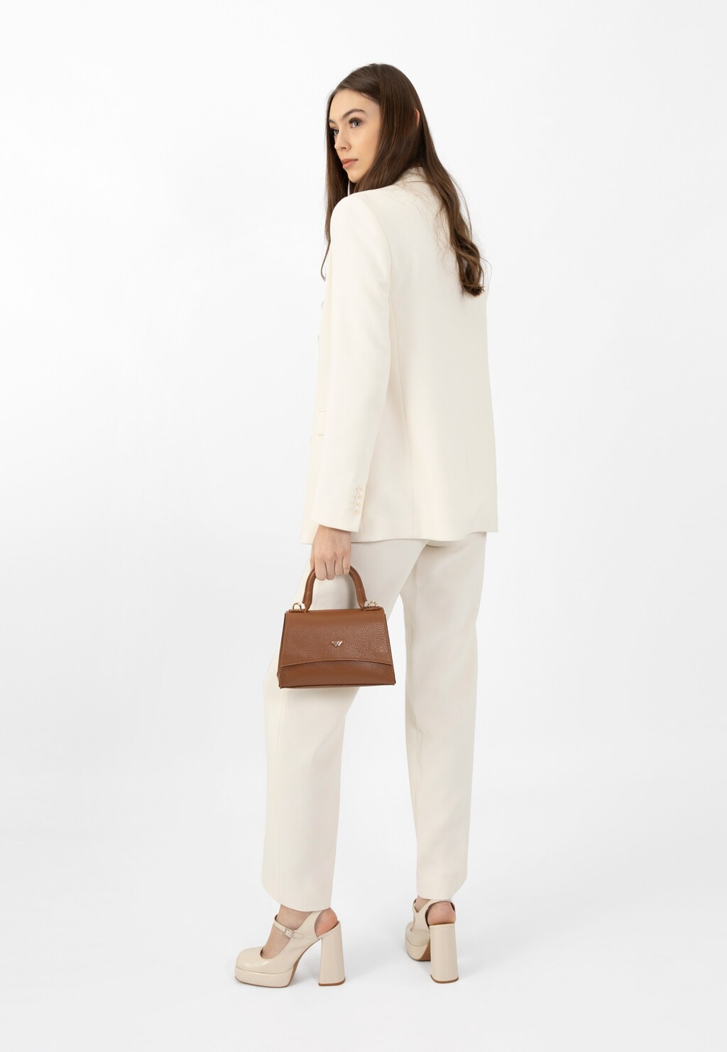 Modelka w białym garniturze prezentująca wysokie beżowe czółenka Wojas 35124-34 na masywnym słupku, trzymająca brązową torebkę, widok z tyłu.