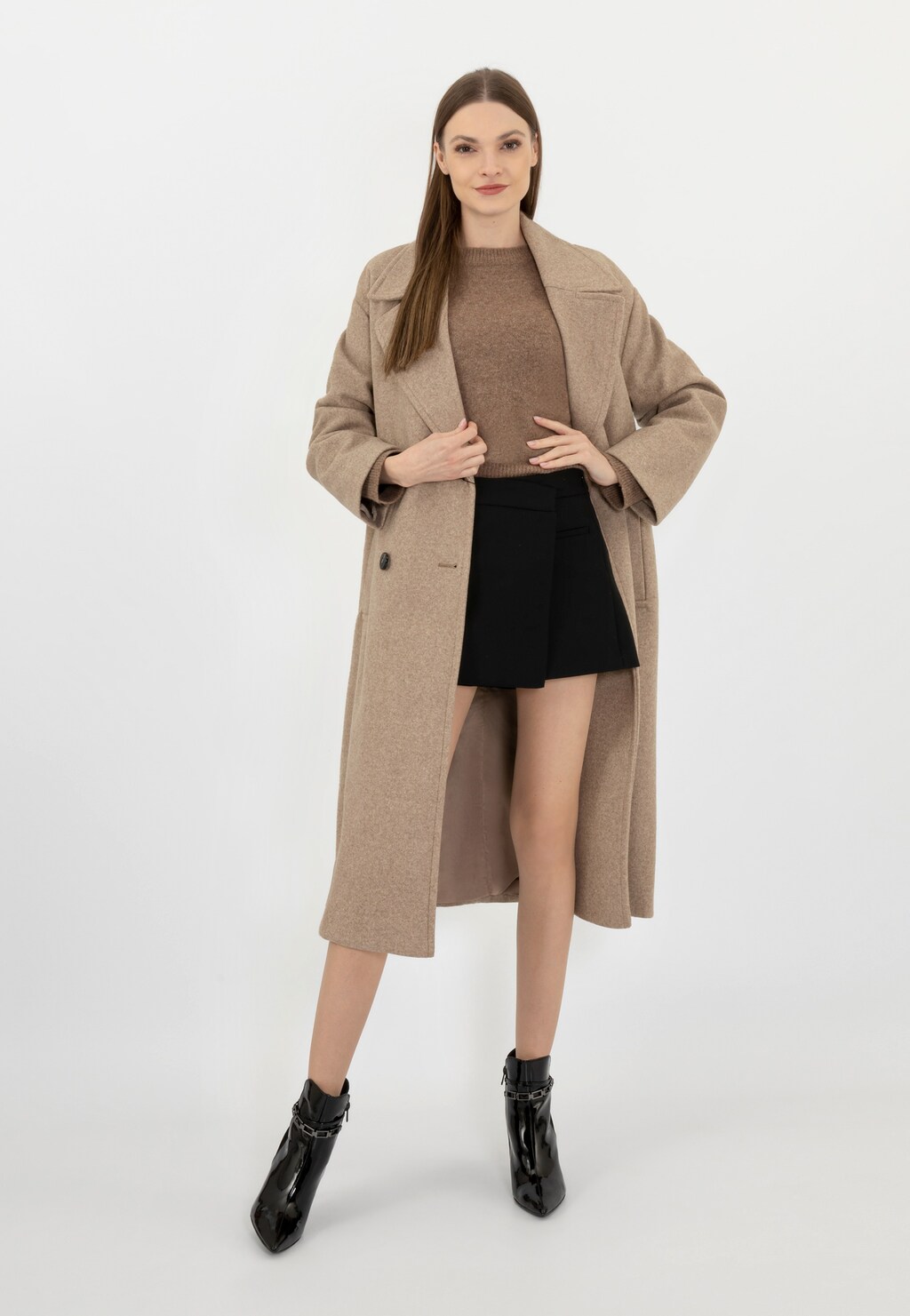 Modelka w beżowym płaszczu i swetrze prezentuje czarne botki Wojas 55209-71 z charakterystycznym łańcuchem wokół kostki. Elegancki, błyszczący model doskonały do stylizacji z krótką spódnicą.