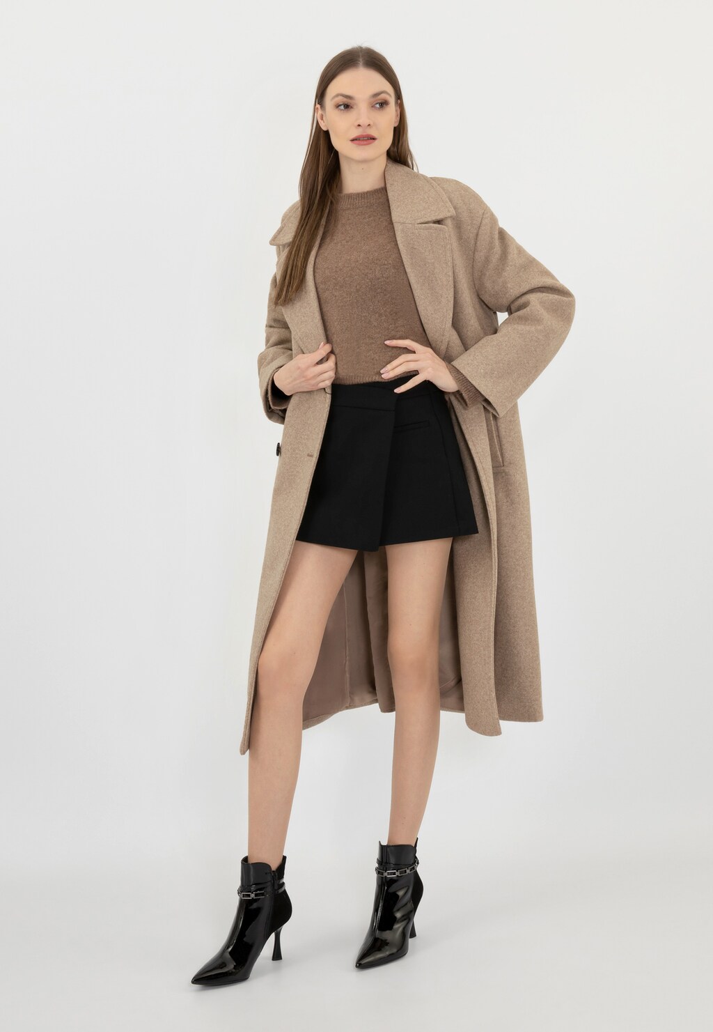 Stylizacja z czarnymi eleganckimi botkami Wojas model 55209-71 z ozdobnym łańcuchem wokół kostki, beżowym płaszczem, brązowym swetrem i czarną mini spódnicą.