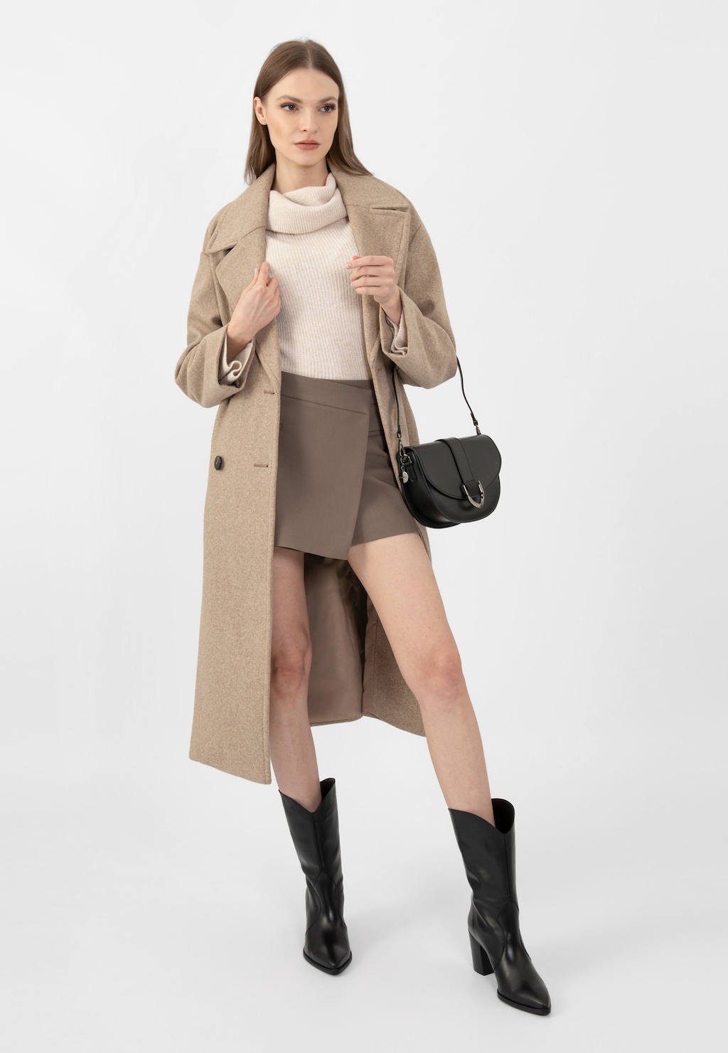 Modelka w beżowym płaszczu i białym golfie prezentuje czarne skórzane kowbojki Wojas model 55239-51 na wysokim obcasie, noszone z brązową spódnicą i czarną torebką.