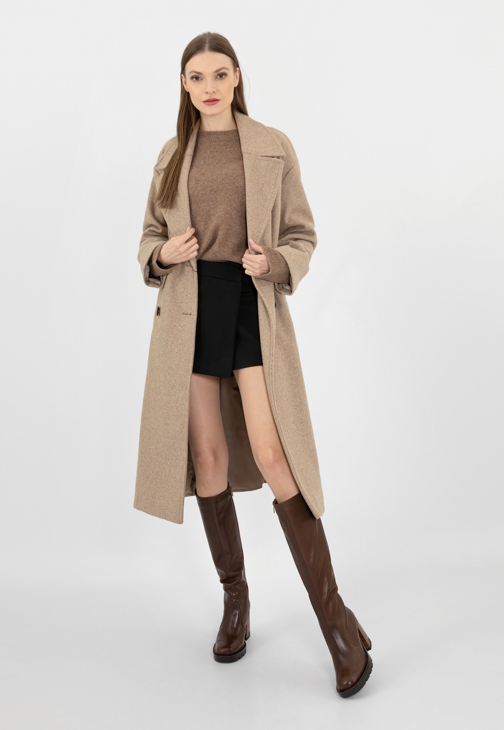 Modelka w stylizacji z beżowym płaszczem, brązowym swetrem i czarną mini spódnicą prezentuje brązowe kozaki damskie Wojas ze skóry licowej model 71050-52 sięgające za kolana.