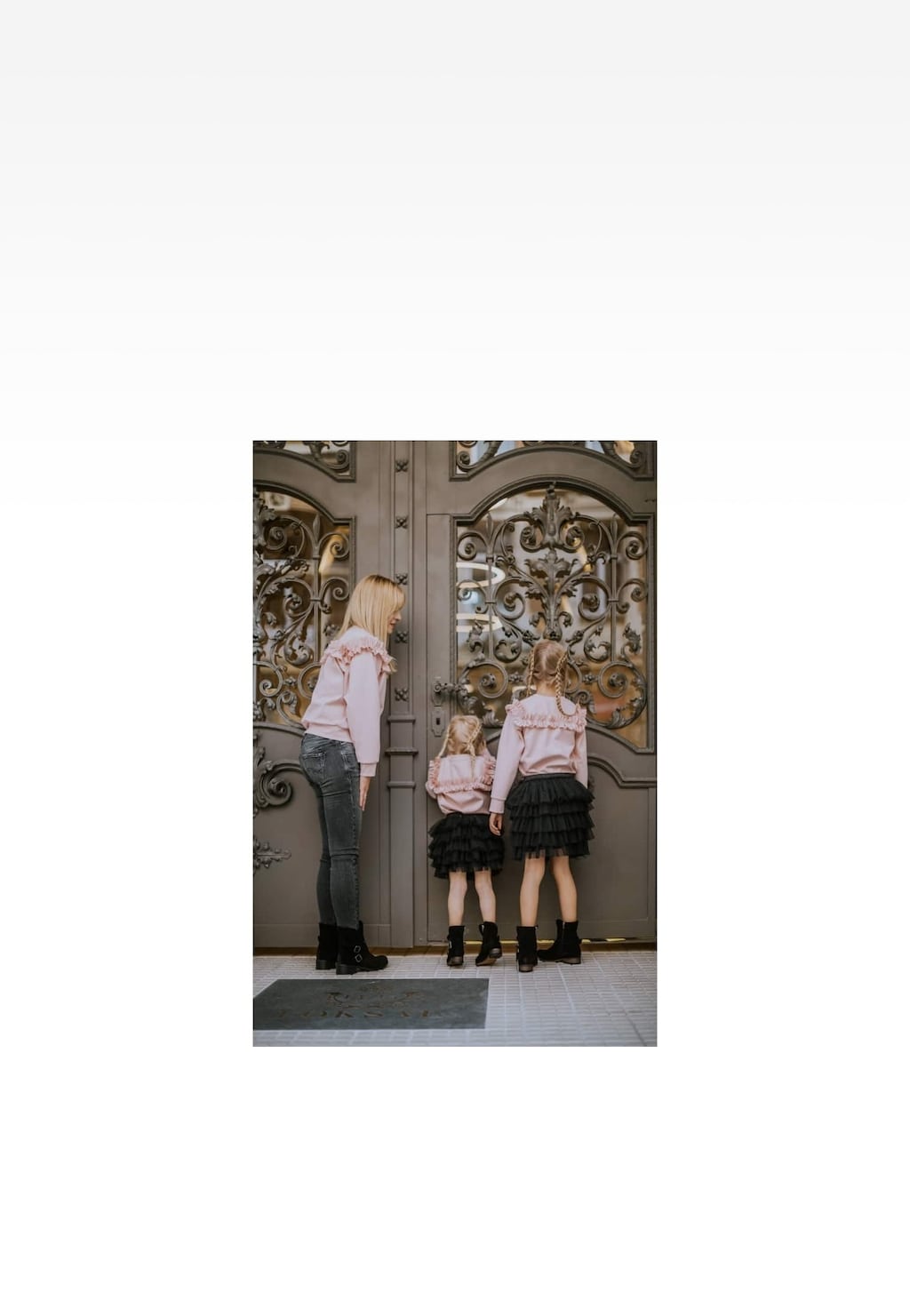 Kobieta z dwiema dziewczynkami w różowych bluzkach i czarnych spódniczkach, stojące przed ozdobnymi drzwiami. Dziewczynki prezentują Botki Wojas x Bartek 8440461 w czarnym kolorze.
