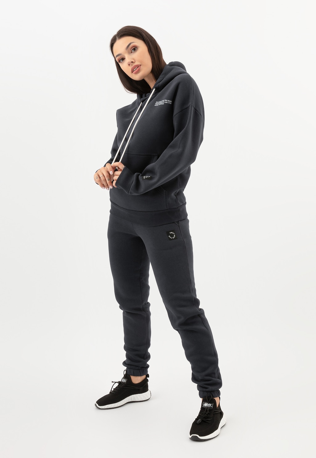 Modelka prezentuje popielate spodnie dresowe RELAKS model R98011-11 w komplecie z bluzą z kapturem, tworząc sportowy zestaw uzupełniony czarnymi butami sportowymi marki Relaks.