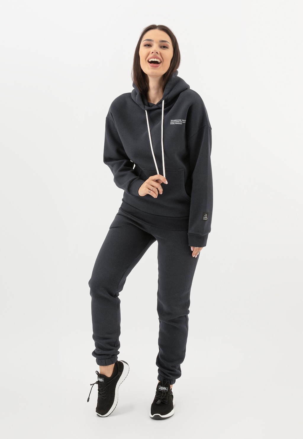 Modelka w popielatym komplecie dresowym RELAKS - bluza z kapturem i spodnie model R98011-11, prezentuje sportową stylizację uzupełnioną czarnymi butami sportowymi.