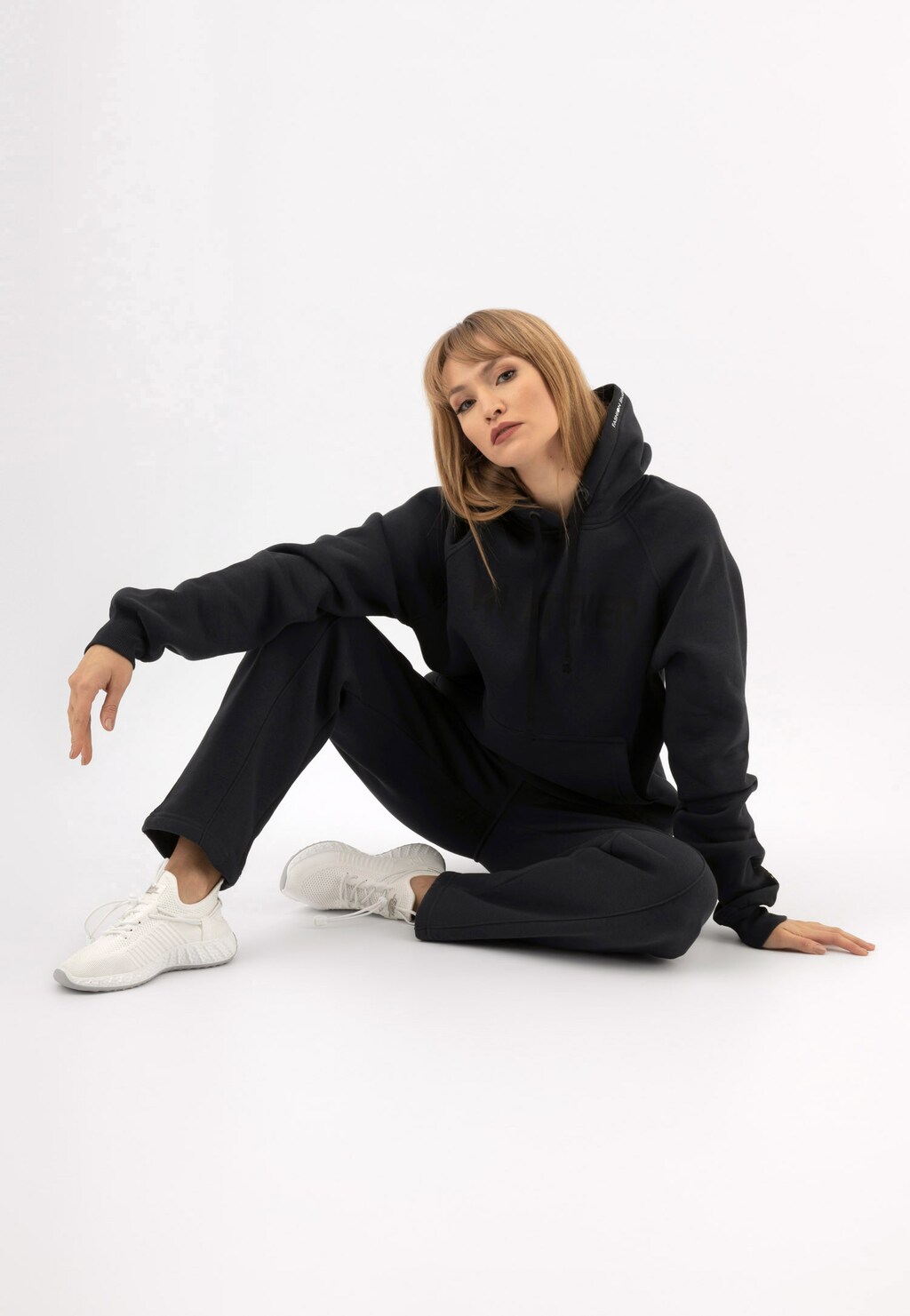 Modelka w ciemnopopielatej bluzie z czarnym napisem RELAKS model R98012-11, prezentowana w wygodnej, siedzącej pozie, ubrana w komplet dresowy z białymi butami marki Relaks.