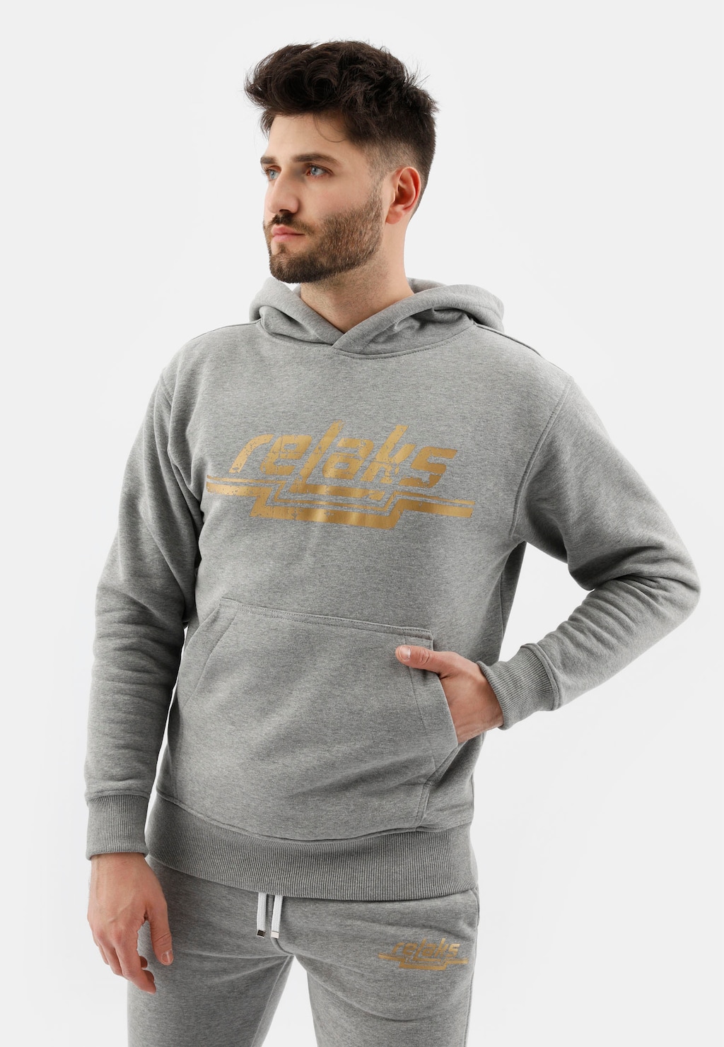 Popielata męska bluza z kapturem RELAKS model 98020-80 ze złotym logo na przodzie. Widoczna kieszeń kangurka, w której model trzyma rękę. Strój uzupełniają spodnie dresowe.
