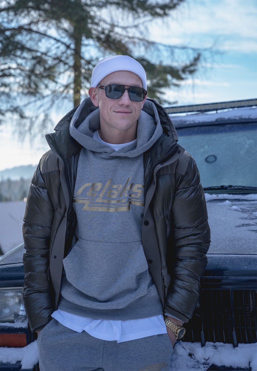 Popielata bluza z kapturem RELAKS model 98020-80 prezentowana w zimowej scenerii, widoczna pod czarną kurtką puchową, uzupełniona białą czapką i okularami przeciwsłonecznymi.