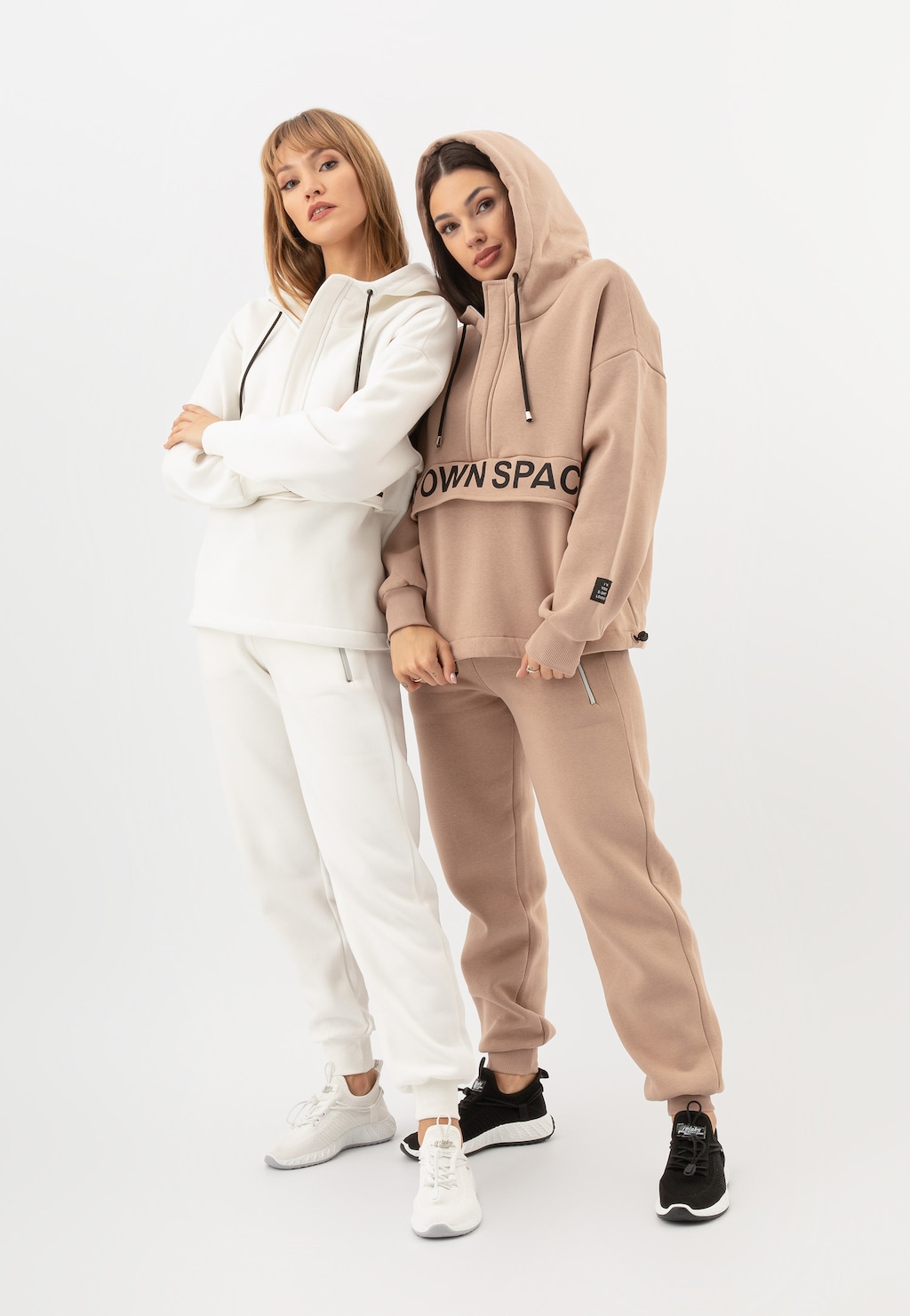 Dwie modelki w dresach - jedna w białym komplecie, druga w beżowych spodniach marki Relaks model R98015-14 z dopasowaną bluzą z kapturem i napisem 