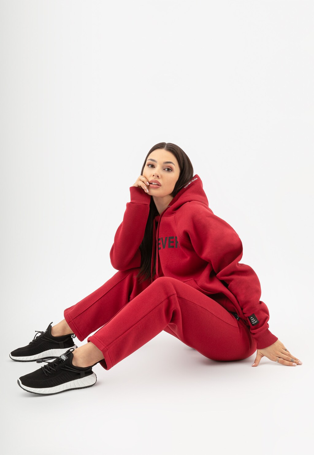Czerwone spodnie dresowe RELAKS R98013-15 o prostym kroju prezentowane na modelce, która siedzi w komplecie z czerwoną bluzą z kapturem i czarnymi butami sportowymi.