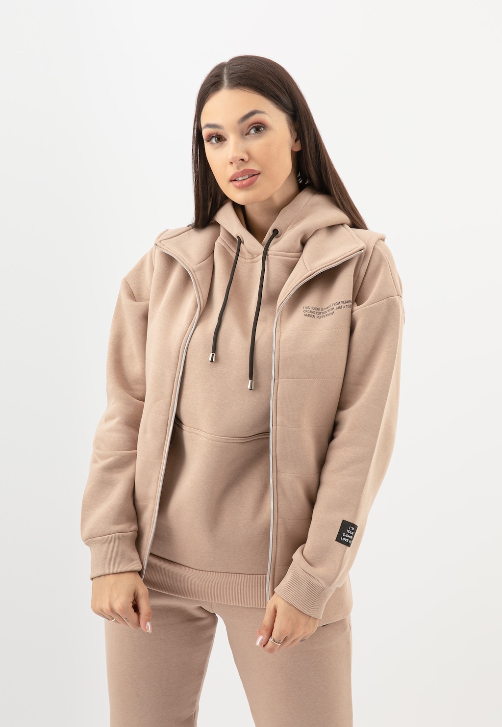 Beżowa dresowa kamizelka damska Relaks R98020-14 prezentowana z rozpiętym zamkiem, noszona na dopasowanej bluzie z kapturem, tworząca komplet z dresowymi spodniami w tym samym kolorze.
