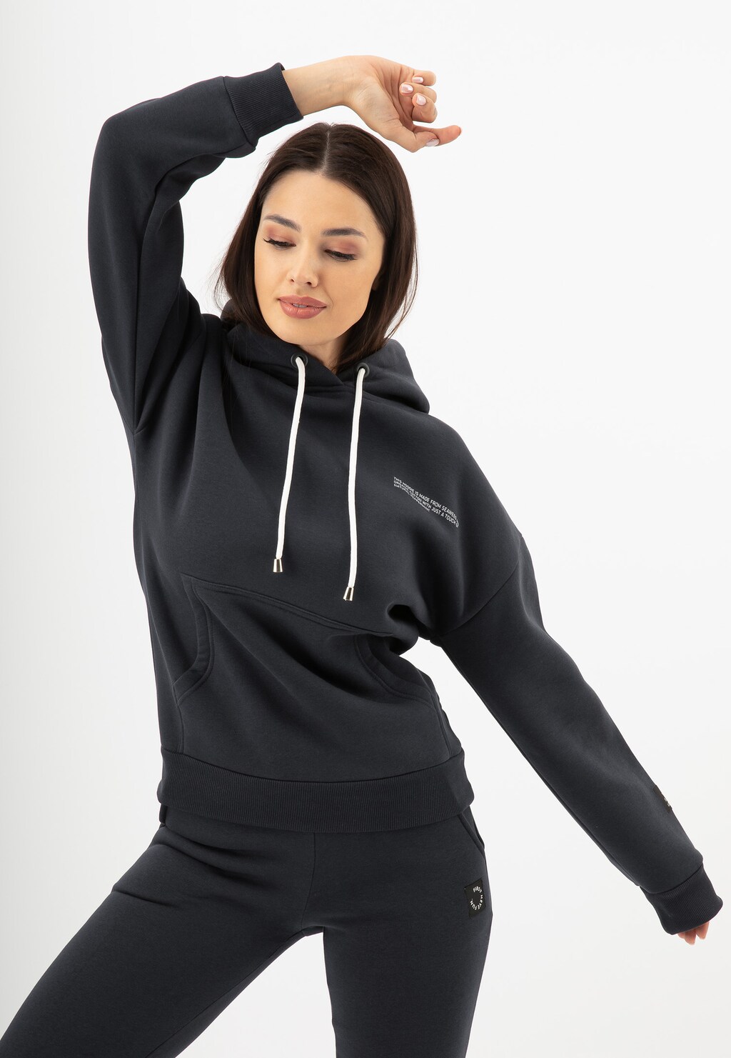 Modelka prezentuje bluzę damską Relaks R98010-11 w kolorze ciemnego popielu z kapturem i białymi sznurkami, w dynamicznej pozie z uniesioną ręką, widoczne dopasowane spodnie w tym samym kolorze.