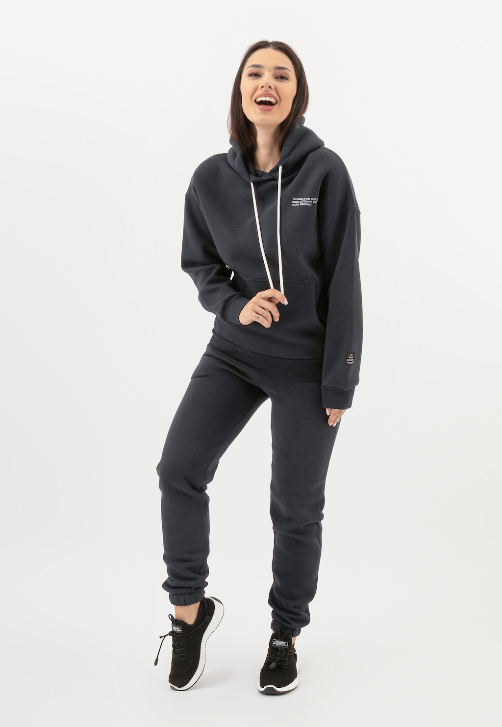 Bluza damska RELAKS model R98010-11 w kolorze ciemnego popielu z kapturem i troczkami, prezentowana w komplecie ze spodniami dresowymi i sportowymi butami.