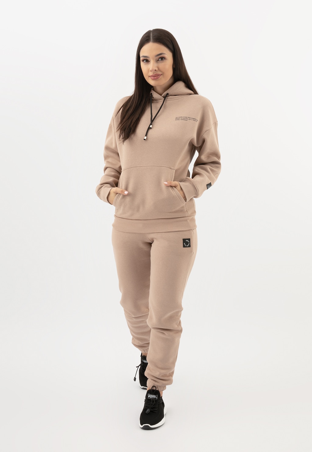 Beżowy dres damski RELAKS model R98017-14 - ciepła bluza z kapturem i kieszenią kangurką oraz dopasowane spodnie, prezentowane na modelce w czarnych butach sportowych.