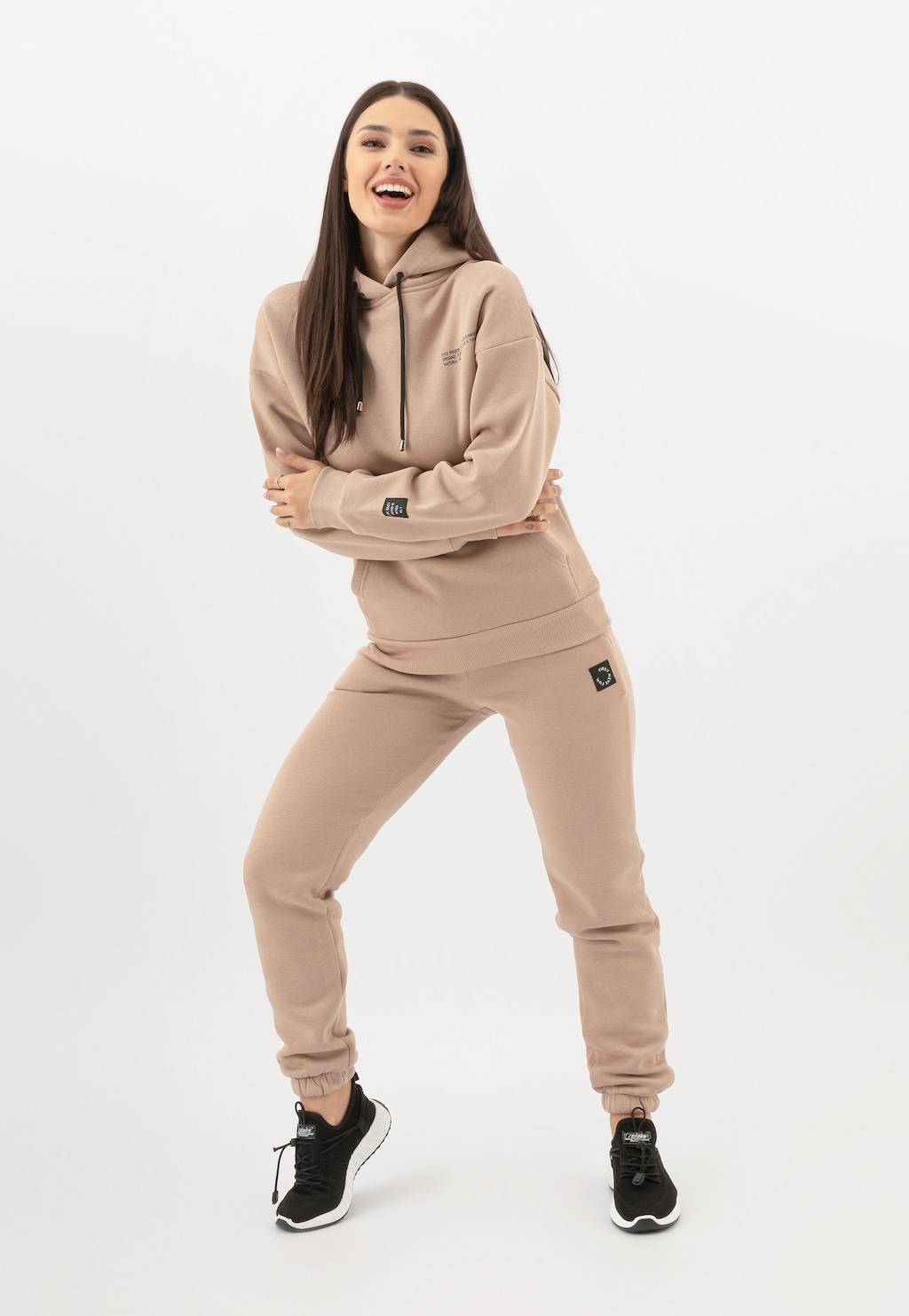 Modelka w komplecie dresowym marki Relaks - beżowe spodnie damskie model R98017-14 i bluza z kapturem w tym samym kolorze, uzupełnione czarnymi butami sportowymi.