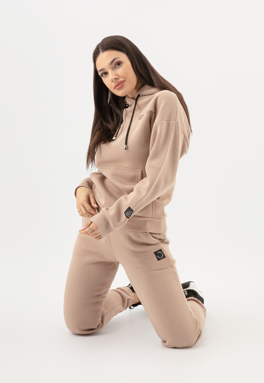 Modelka prezentująca beżowy dres marki Relaks - ciepłe spodnie damskie model R98017-14 z dopasowaną bluzą z kapturem, w pozycji klęczącej.