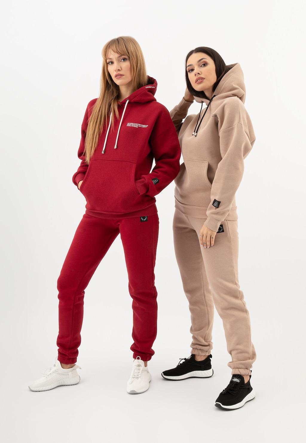 Dwie modelki w dresach - jedna w czerwonym komplecie, druga w beżowych spodniach Relaks R98017-14. Wygodne dresy z kapturem, idealne na chłodniejsze dni.