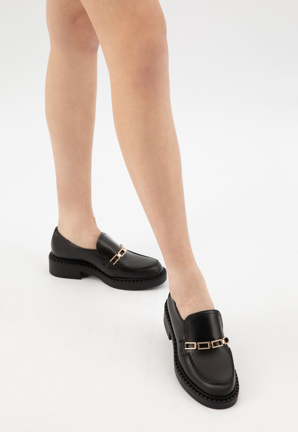 Czarne mokasyny damskie Wojas model 46240-31 ze skóry florentic, prezentowane na nogach modelki. Buty zdobi elegancka złota ozdoba na przodzie.