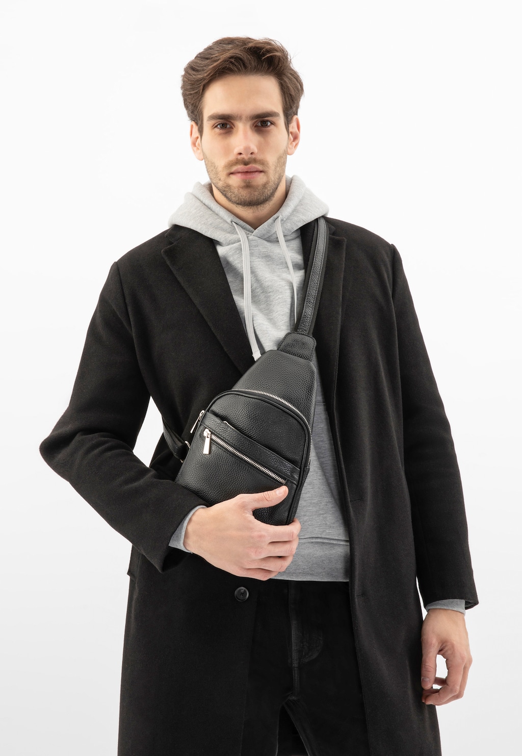 Model w czarnym płaszczu i szarej bluzie prezentuje czarną torbę męską ze skóry licowej Wojas 80193-51, noszoną na ukos przez ramię, z widocznymi kieszeniami na zamek.