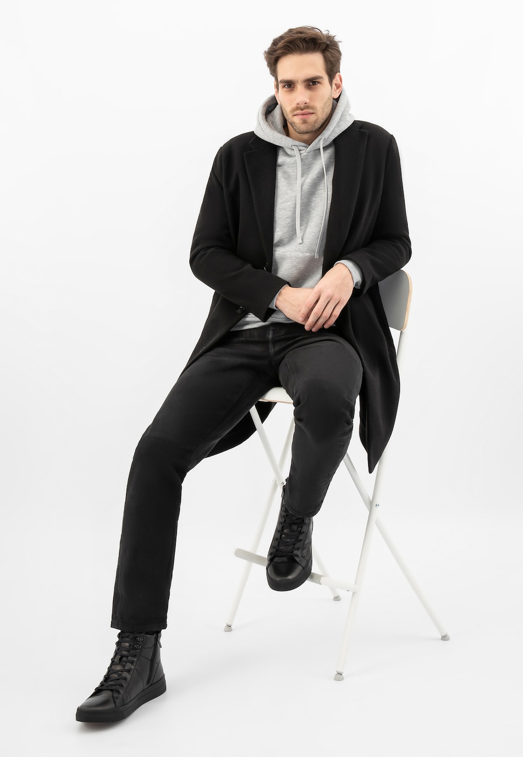 Model siedzący na białym krześle w czarnym płaszczu i szarej bluzie z kapturem, prezentujący czarne trzewiki typu trampki Wojas 24096-51 z futerkowym ociepleniem.