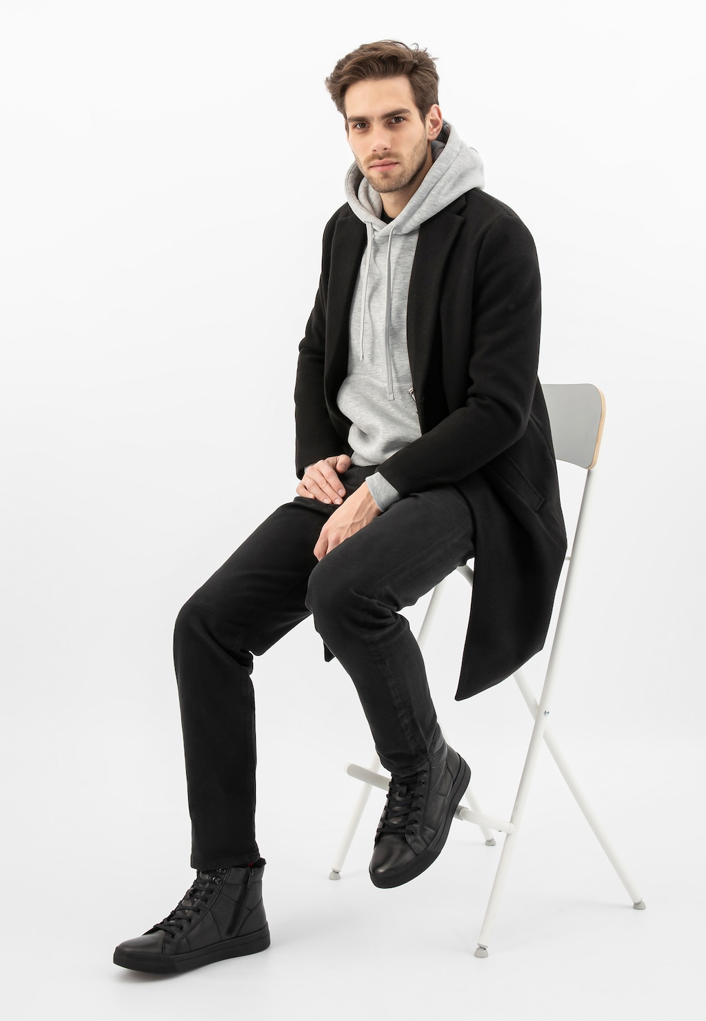 Model w czarnym płaszczu i szarej bluzie z kapturem, prezentujący czarne trzewiki Wojas model 24096-51 z futerkowym ociepleniem, siedzący na białym krześle.