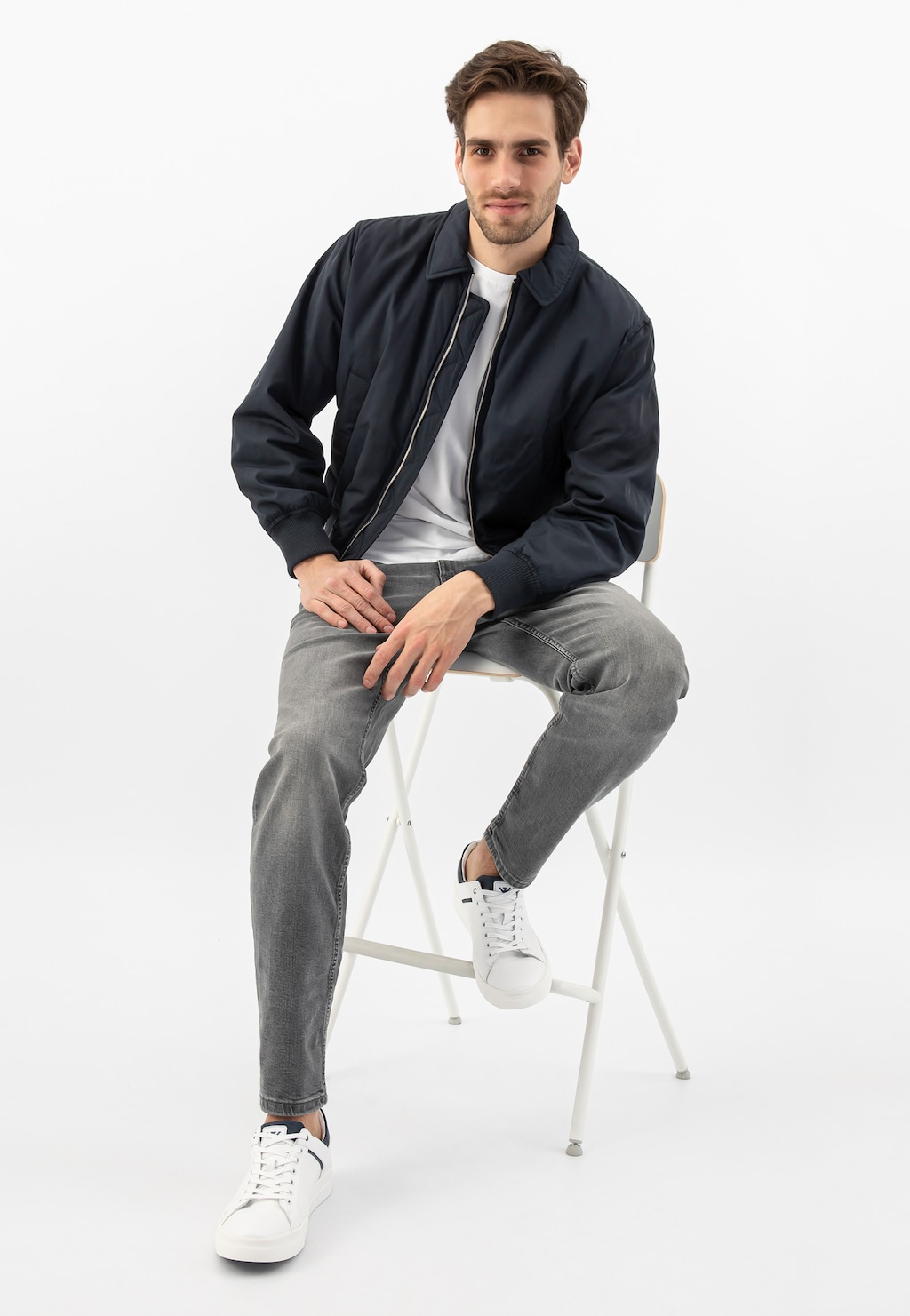 Model w stylizacji casualowej prezentuje białe trampki męskie Wojas 10199-50 z granatowymi wstawkami, siedząc na białym krześle w granatowej kurtce i szarych jeansach.