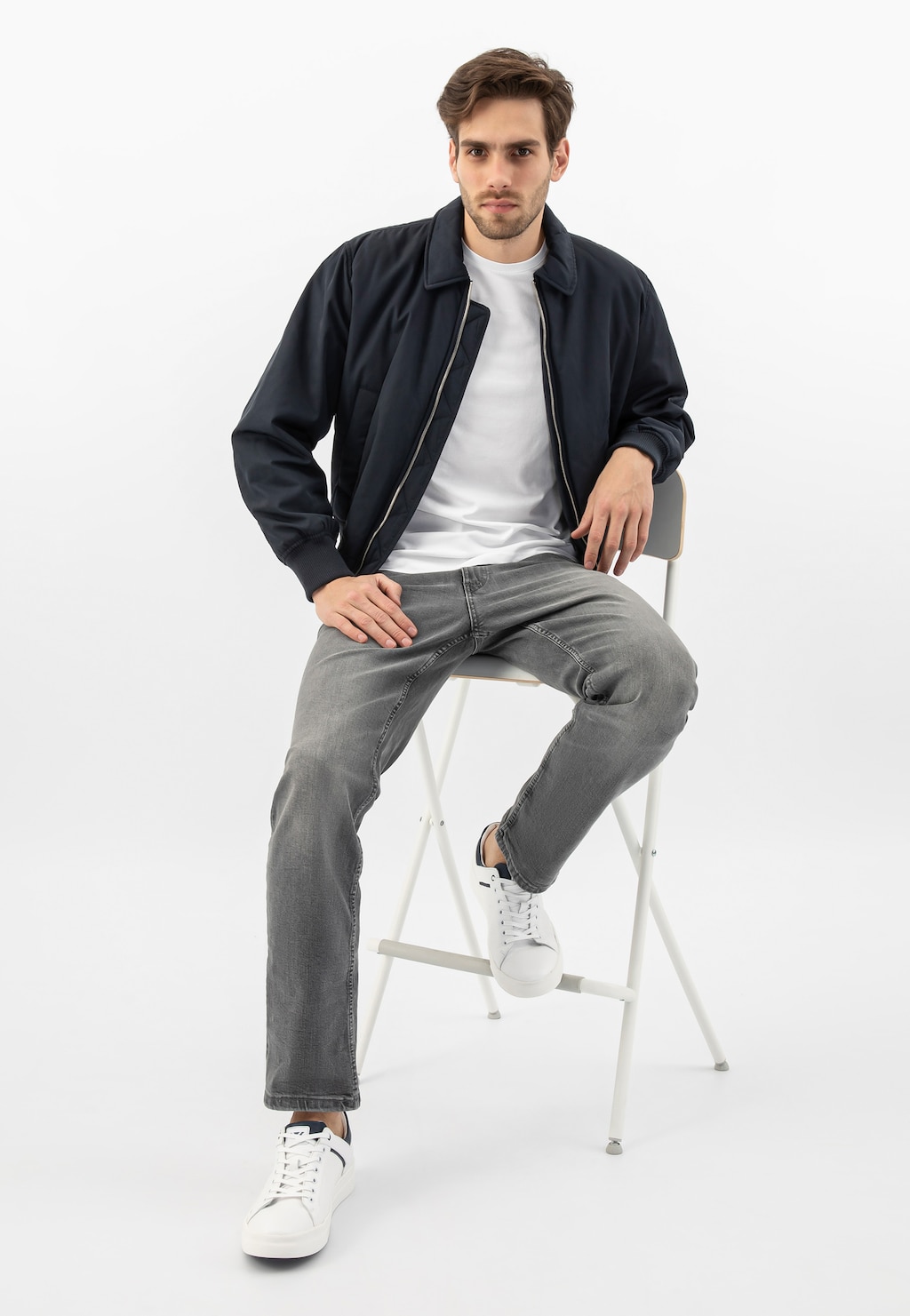 Model siedzący na krześle w stylizacji z granatową kurtką i białymi trampkami Wojas model 10199-50 z granatowymi wstawkami, szarymi jeansami i białym t-shirtem.