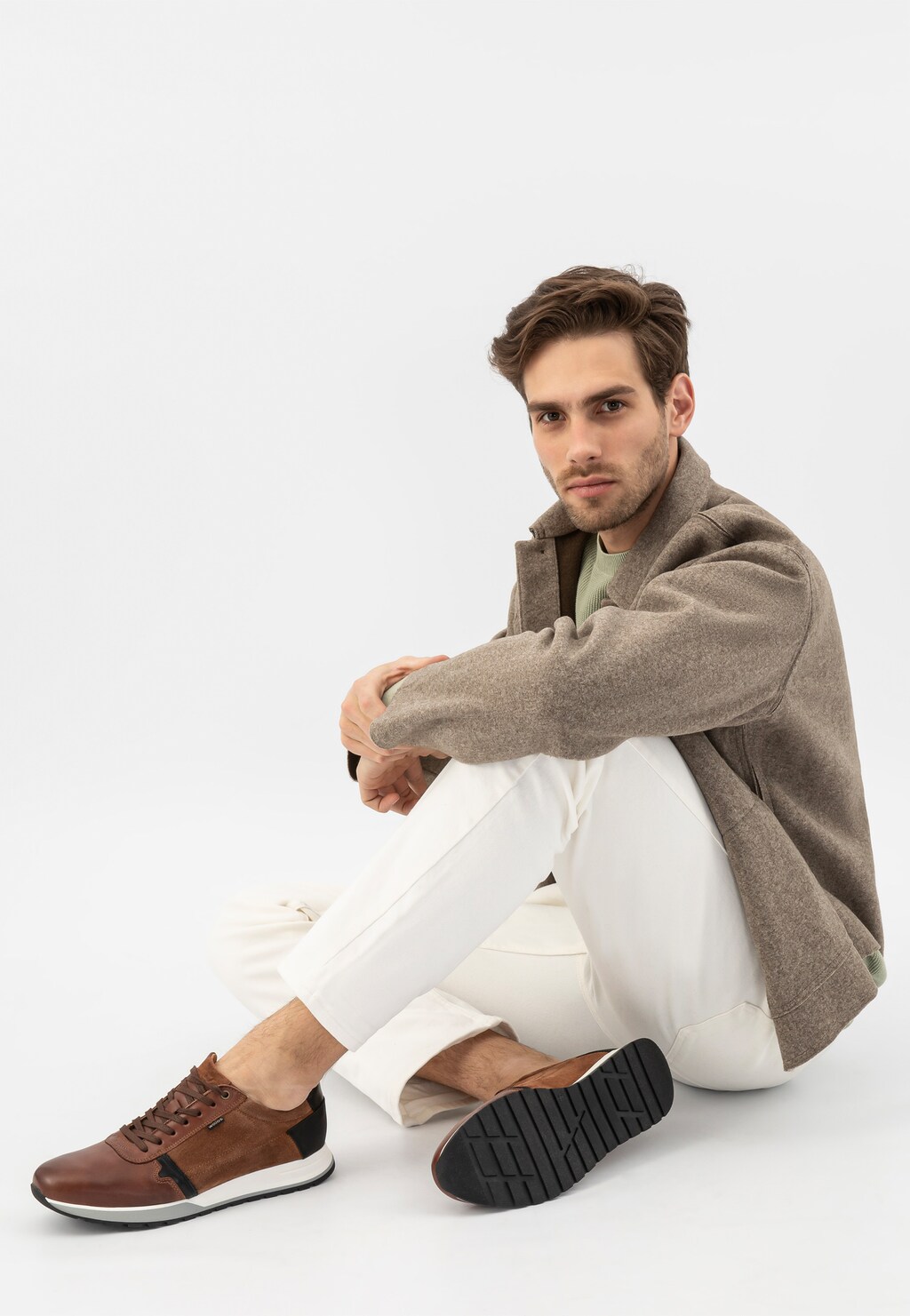 Model w jasnobrązowych sneakersach Wojas 10021-73 z czarnymi elementami, ubrany w szary płaszcz i białe spodnie, siedzący w swobodnej pozie.