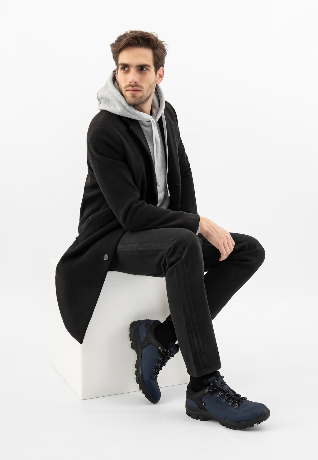 Model w eleganckim czarnym płaszczu i szarej bluzie z kapturem prezentuje granatowo-czarne trekkingi męskie Wojas 9377-80 z dzianiną dystansową, siedząc na białym podeście.