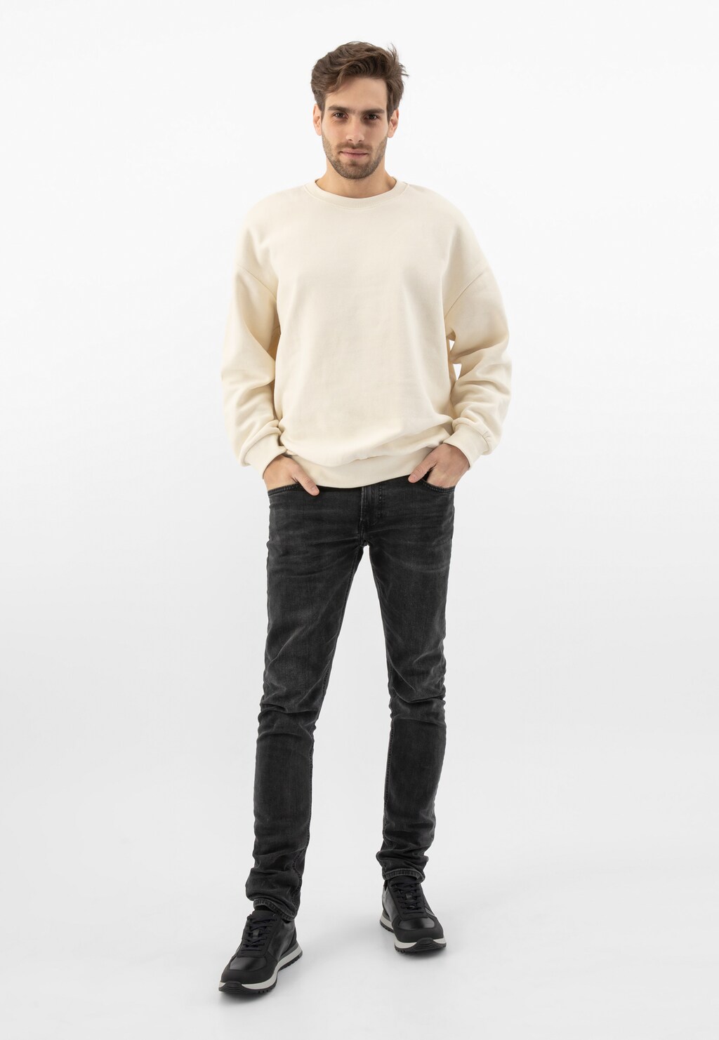 Model w kremowej bluzie i czarnych jeansach prezentuje czarne sneakersy męskie Wojas 10132-71 z szarymi elementami na podeszwie, widoczne na stopach.