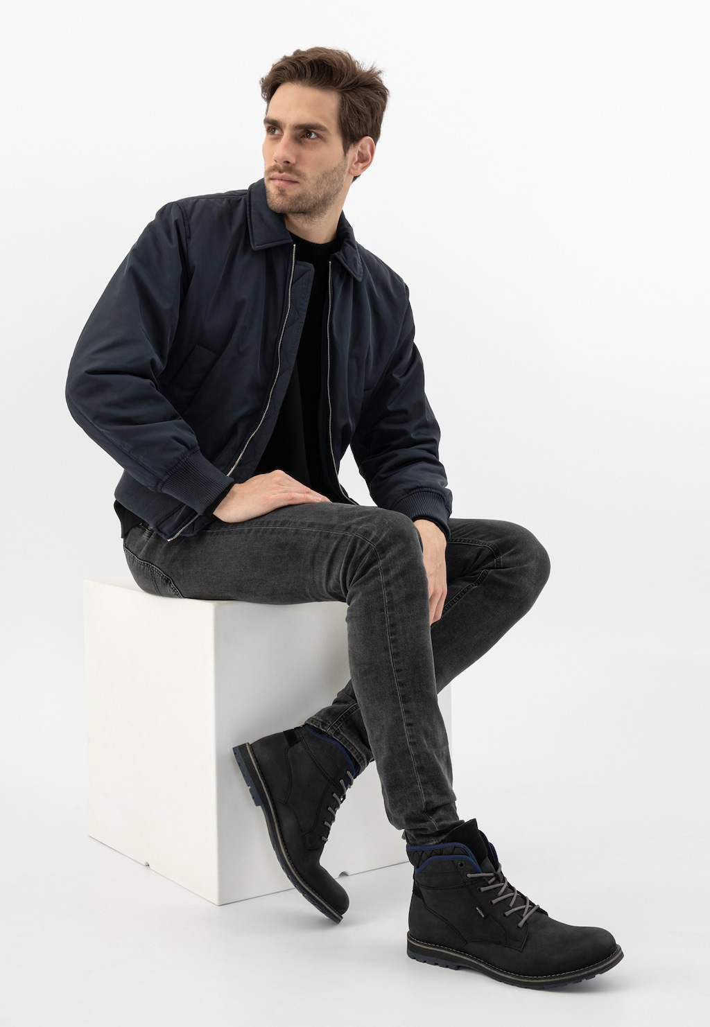 Mężczyzna w granatowej kurtce i czarnych jeansach prezentujący czarne workery Wojas model 24031-71 z charakterystycznymi granatowymi akcentami, siedzący na białym postumencie.