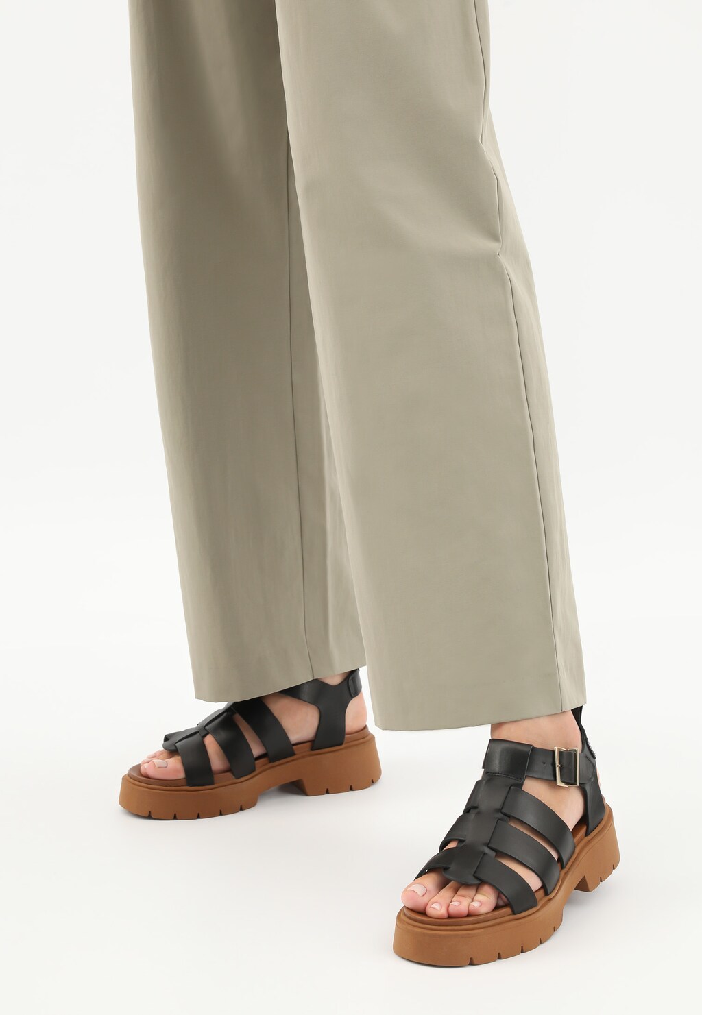 Czarne sandały damskie Wojas model 76136-51 z szerokimi paskami na brązowej, masywnej podeszwie, zapinane na klamrę, noszone do beżowych szerokich spodni.