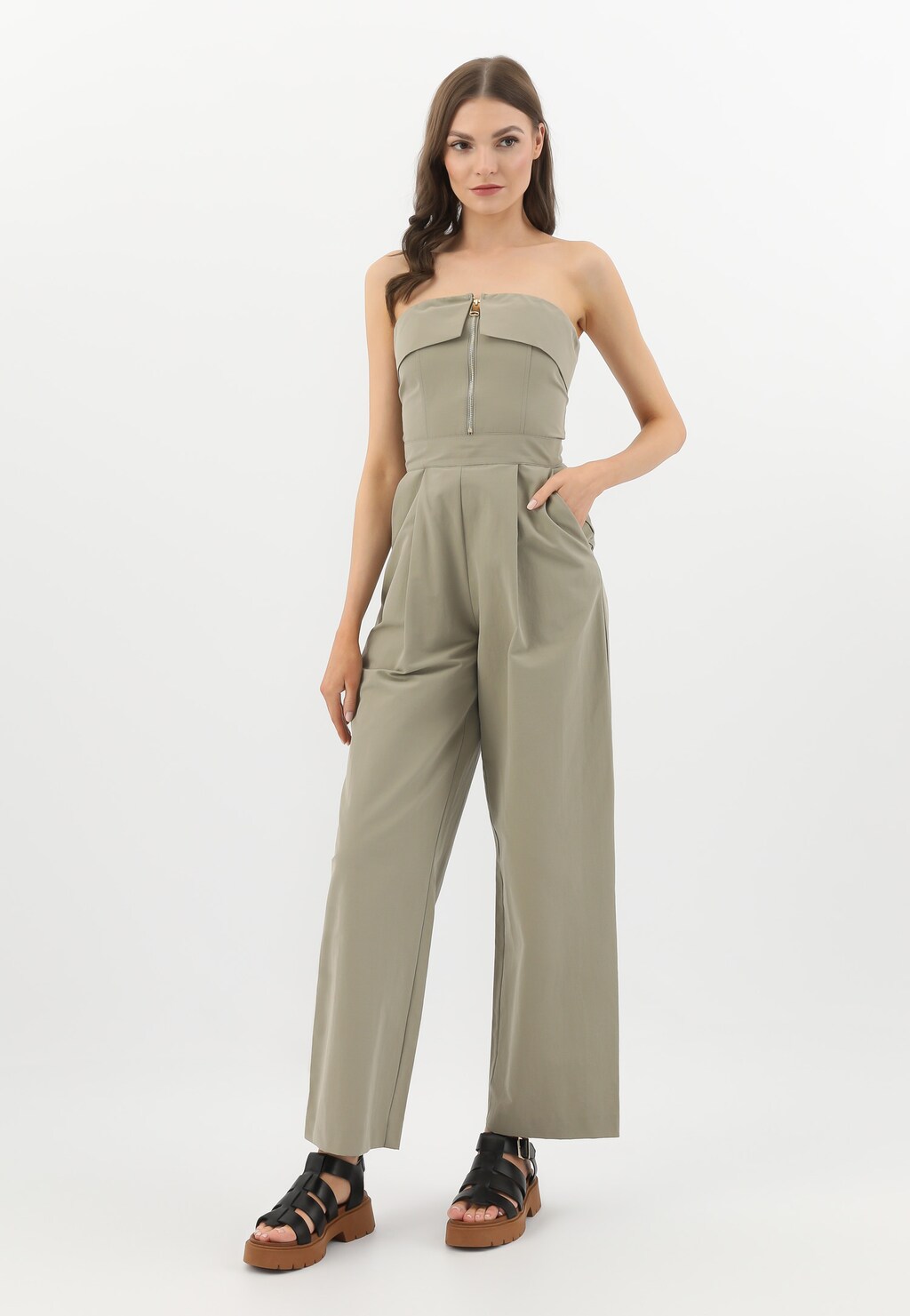 Modelka w beżowym kombinezonie prezentuje czarne sandały damskie Wojas model 76136-51 z szerokimi paskami na brązowej platformie, widoczne z przodu.