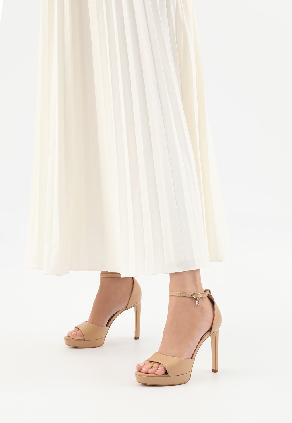 Eleganckie beżowe sandały Wojas model 76152-50 na wysokiej szpilce z platformą, widoczne na nogach osoby w białej plisowanej spódnicy. Pasek przy kostce z ozdobną zawieszką.