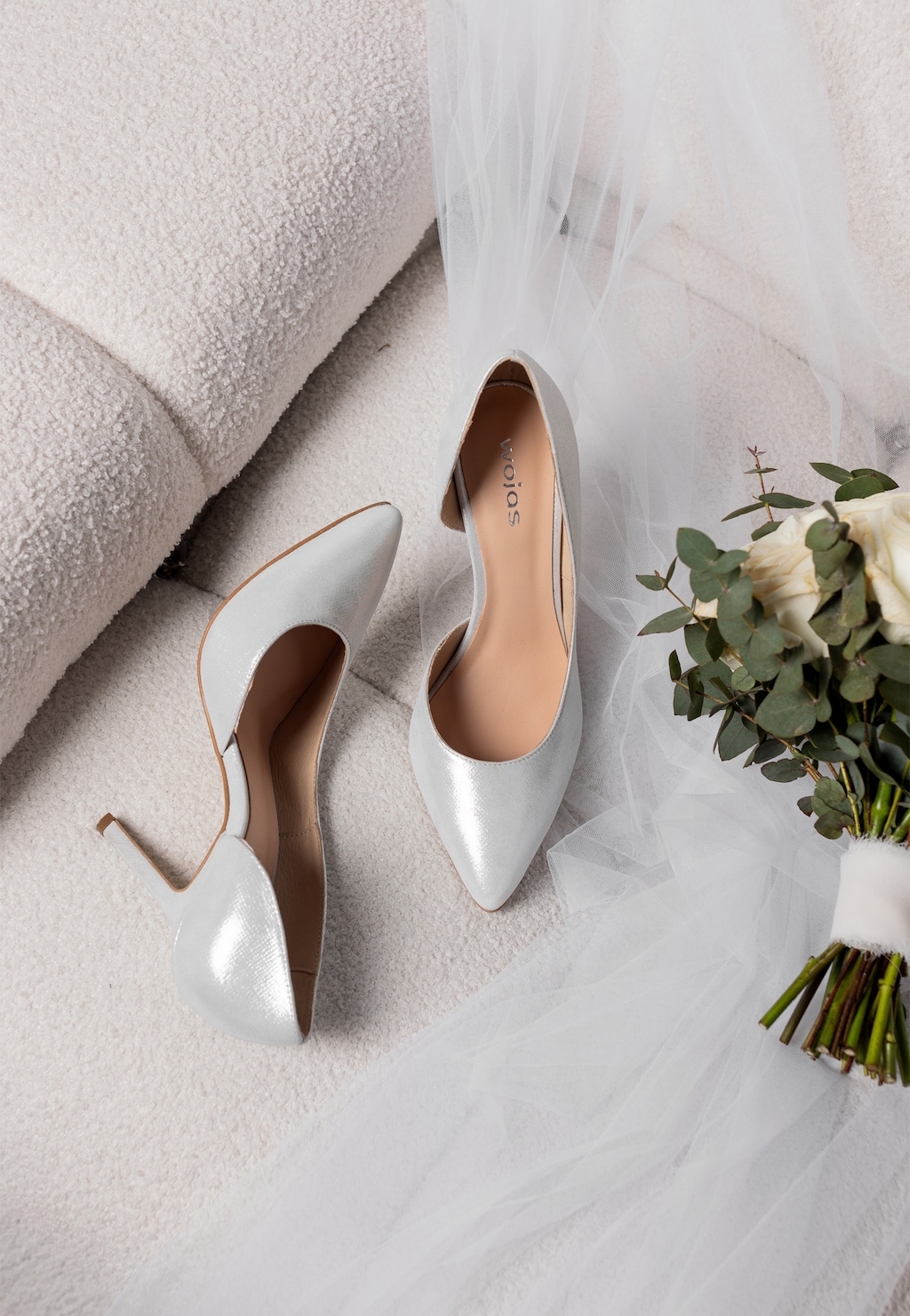 Srebrne eleganckie czółenka Wojas model 35105-59 na szpilce, ułożone na delikatnym welonie ślubnym, obok bukietu białych róż z eukaliptusem.