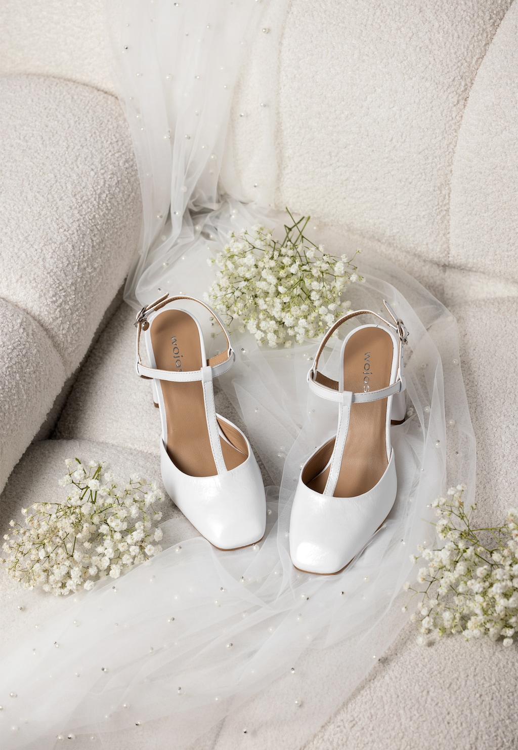 Białe czółenka Wojas model 35152-59 na słupku z odkrytą piętą, ułożone na delikatnym tiulu, otoczone białymi kwiatami gipsówki, w romantycznej ślubnej aranżacji.