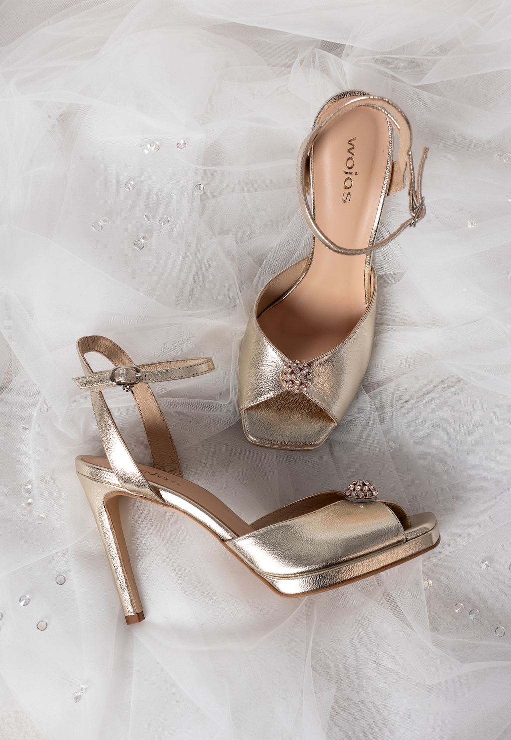 Złote sandały Wojas model 76187-58 na wysokiej szpilce z elegancką kryształkową ozdobą na przodzie, zapinane na pasek, ułożone na delikatnym tiulu z kryształkami.