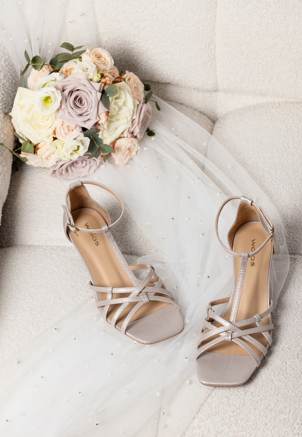 Srebrne sandały Wojas model 76189-59 na niskim obcasie z paseczkami, ułożone na welonie ślubnym obok bukietu pastelowych róż w odcieniach białym i różowym.