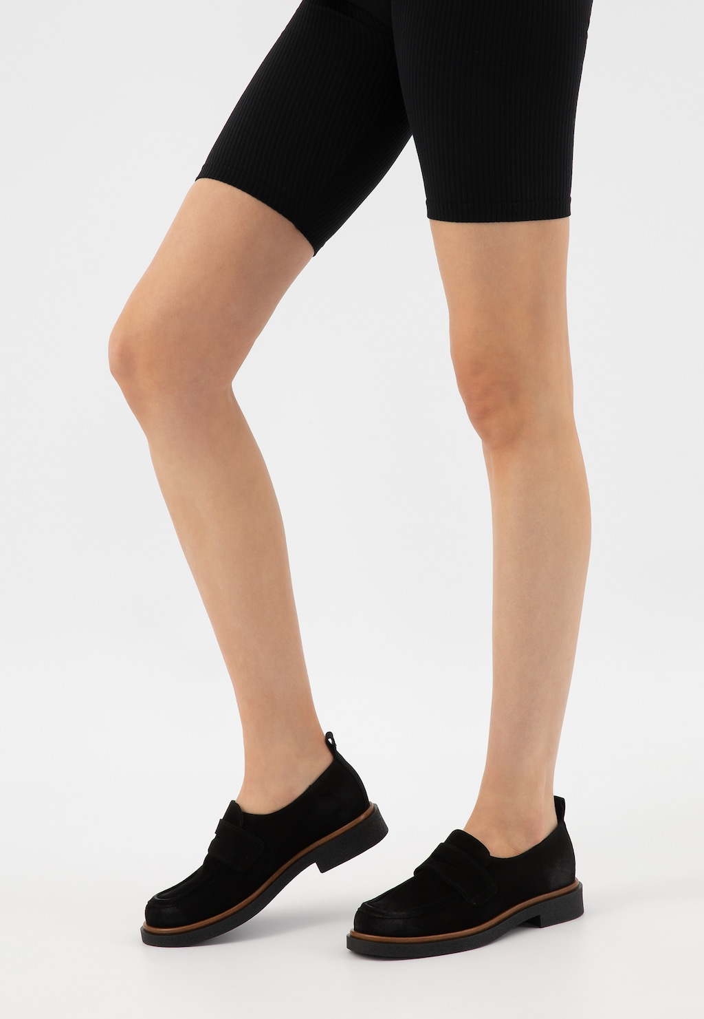 Nogi modelki w czarnych legginsach typu biker, prezentujące czarne mokasyny damskie Wojas penny loafers model 46279-61 w stylu casual z brązowym obramowaniem podeszwy.