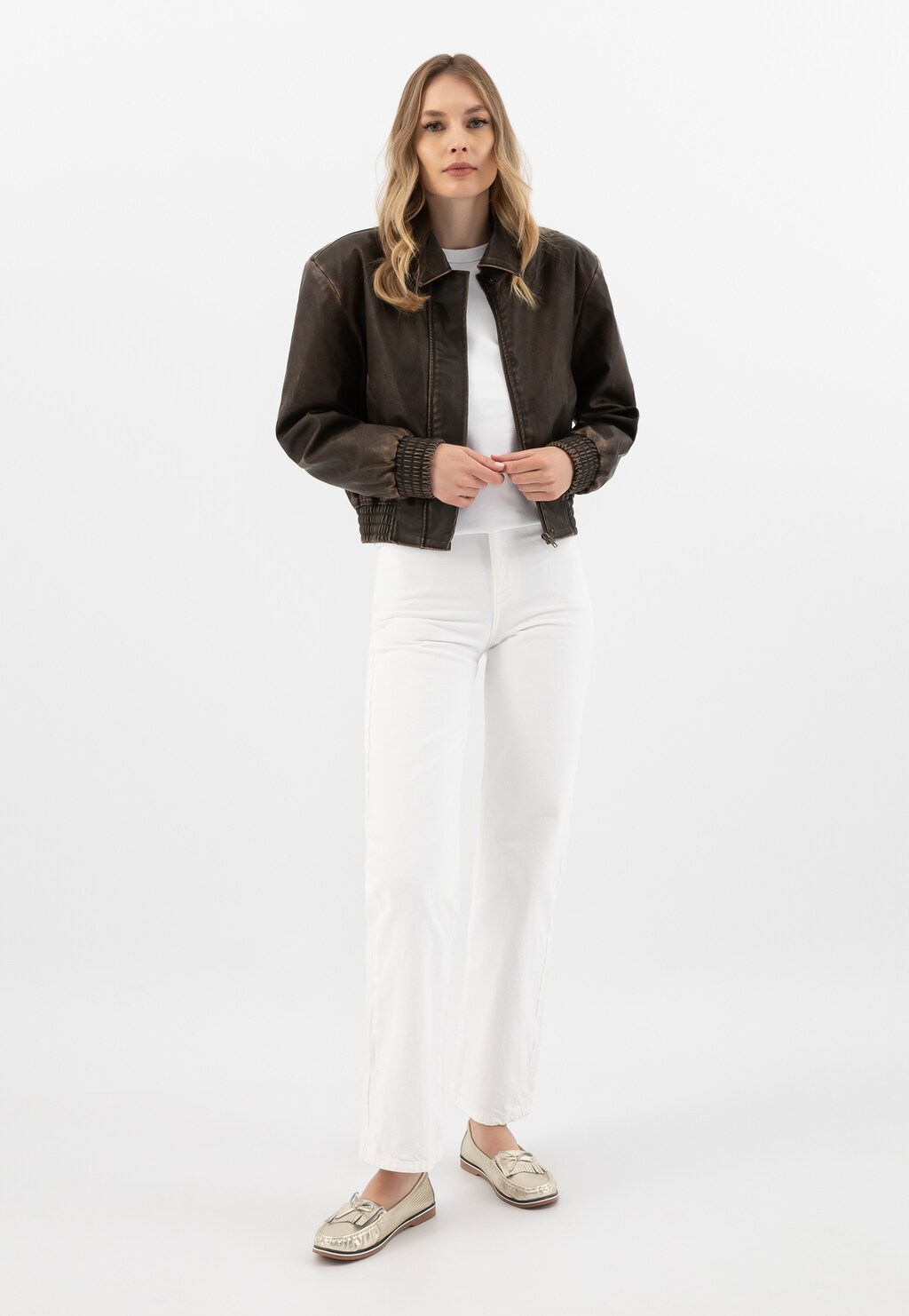 Modelka w brązowej kurtce i białym stroju prezentuje złote mokasyny Wojas model 46293-58 na gumowej podeszwie, idealne do codziennych stylizacji.