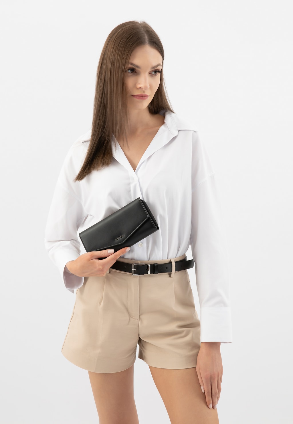 Osoba w białej koszuli i beżowych szortach prezentująca czarny skórzany portfel damski Wojas model 91089-51, trzymany w dłoni na tle eleganckiej stylizacji.