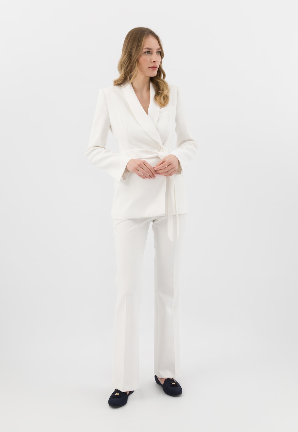 Elegancki biały garnitur damski z marynarką z wiązaniem, uzupełniony granatowymi loafersami Wojas model 45001-67 z ozdobnymi chwostami.