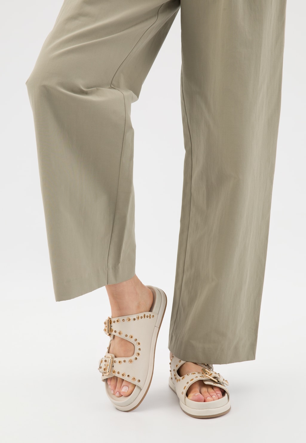 Beżowe płaskie klapki skórzane Wojas model 74125-54 ze złotymi nitami i klamrami, noszone do eleganckich beżowych spodni, widok z perspektywy frontalnej.