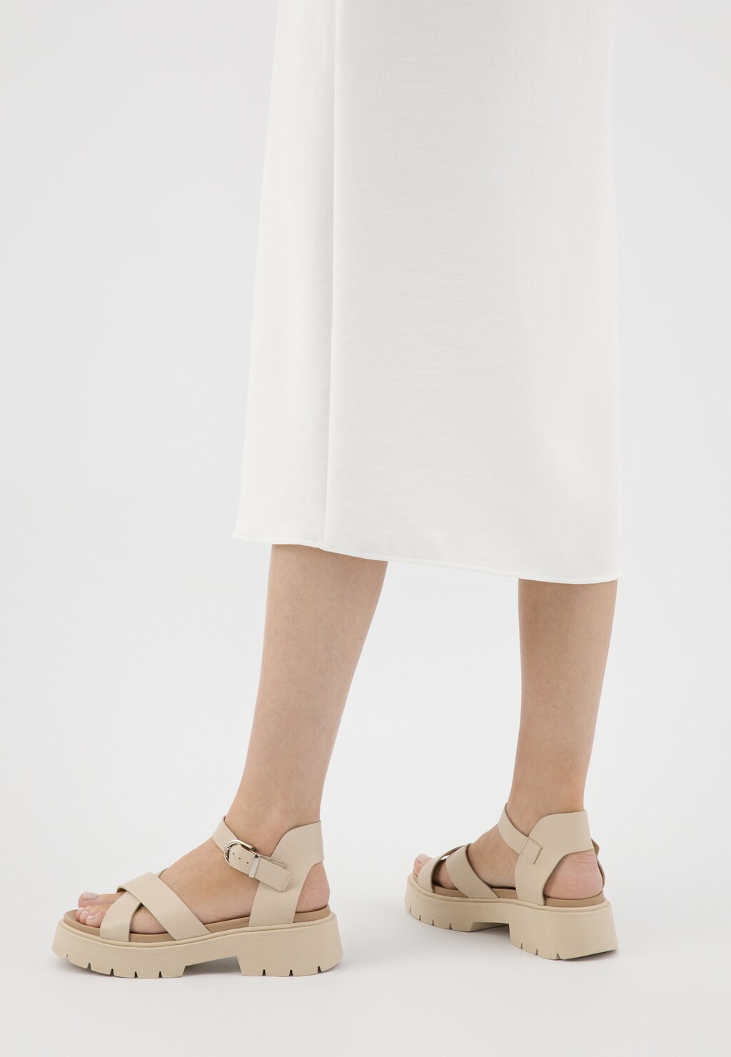 Beżowe sandały Wojas model 76135-54 z masywnym obcasem i grubą podeszwą, zapinane na pasek. Widok z tyłu na nogi modelki w białej spódnicy.