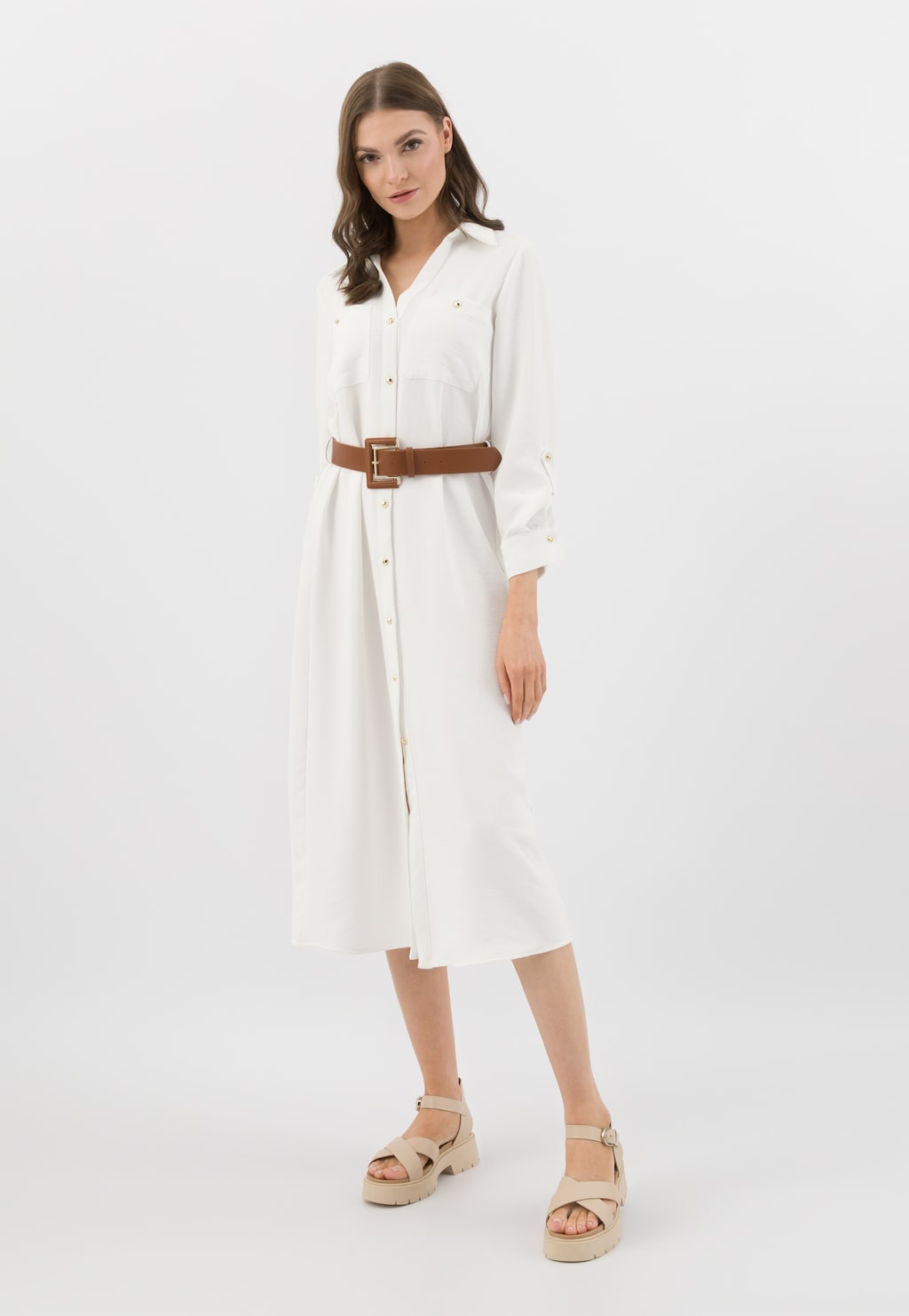 Modelka w białej sukience koszulowej z brązowym paskiem, prezentująca beżowe sandały Wojas na masywnym obcasie model 76135-54, idealne uzupełnienie eleganckiej stylizacji.