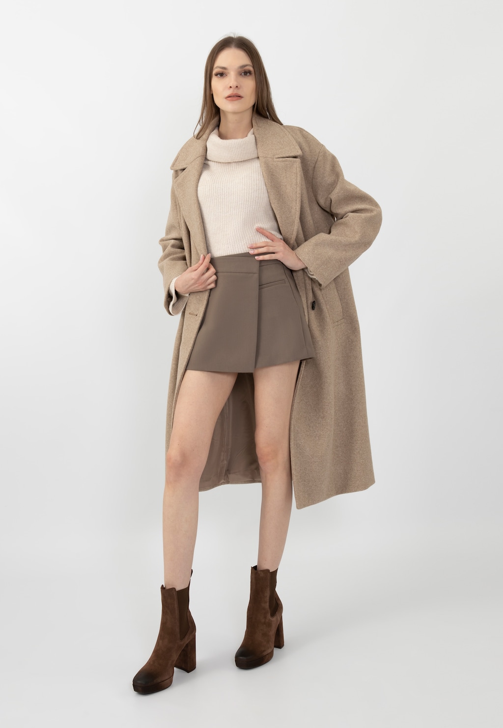 Modelka w eleganckim beżowym płaszczu i krótkiej spódnicy, prezentująca brązowe sztyblety damskie Wojas na wysokim słupku model 55212-62, idealne do jesiennych stylizacji.