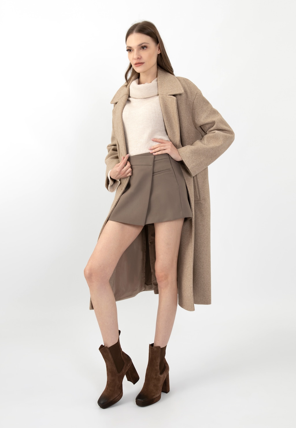 Modelka w eleganckim beżowym płaszczu i krótkiej spódnicy, prezentująca brązowe sztyblety damskie Wojas na wysokim słupku model 55212-62, widoczne w pełnej okazałości.