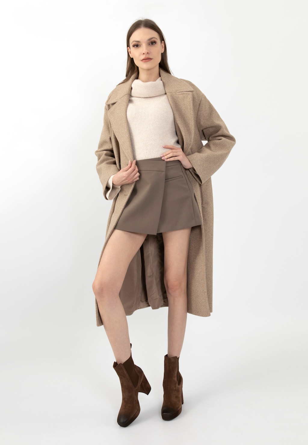 Modelka w jesiennej stylizacji prezentuje brązowe sztyblety damskie Wojas na wysokim słupku model 55212-62, zestawione z beżowym płaszczem, jasnym golfem i brązową mini spódnicą.