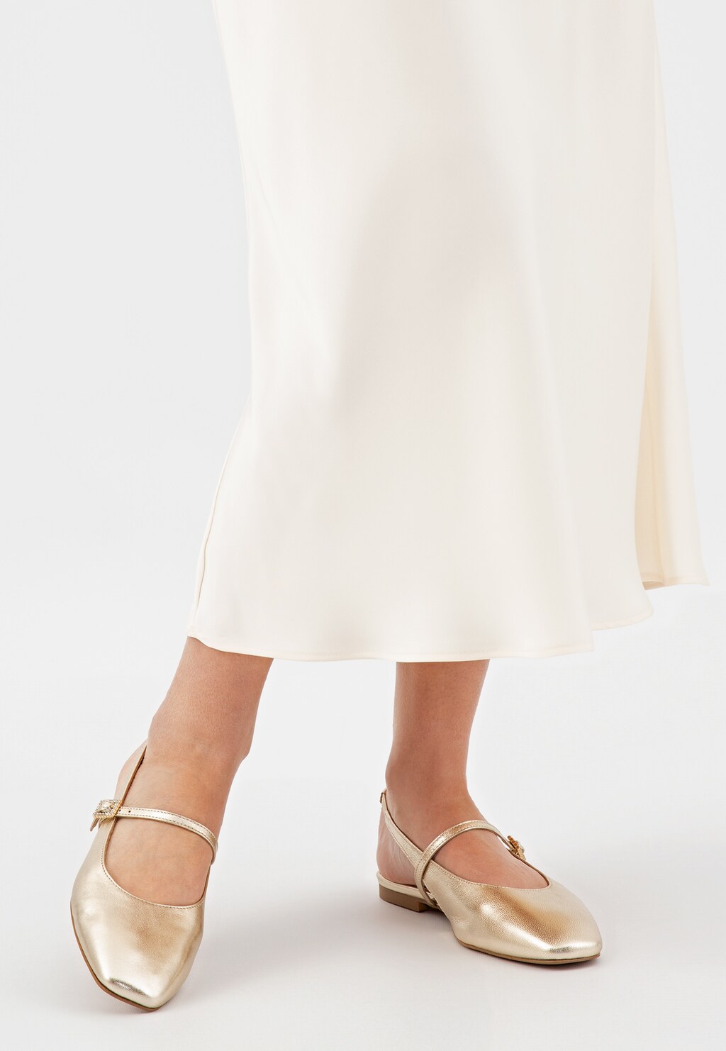 Złote baleriny Wojas model 44047-58 z odkrytą piętą widoczne na nogach osoby w kremowej spódnicy. Eleganckie obuwie z paskiem zapinającym na kostce.